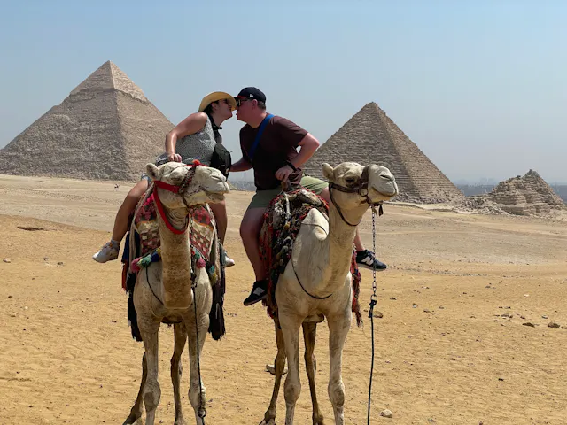 Two people on camels in front of the Egyptian pyramids in the desert.