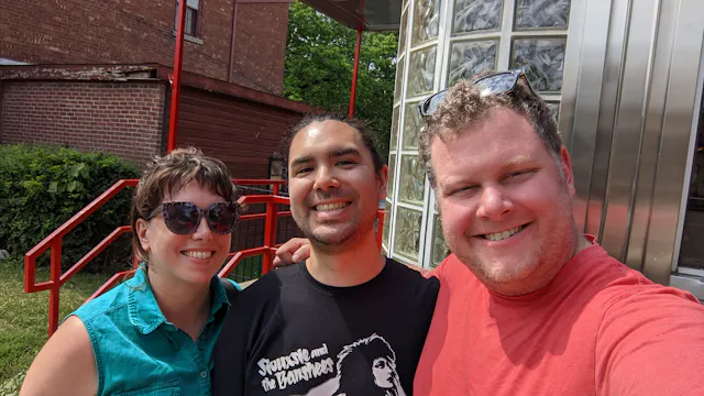 Three men posing together outdoors in front of a brick building with metal railings.