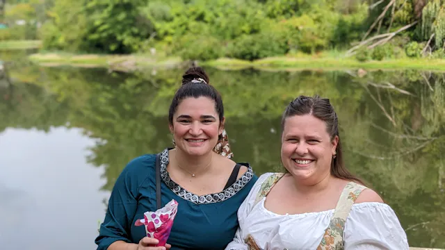 Two women smiling by a river or water body with green foliage behind them.
