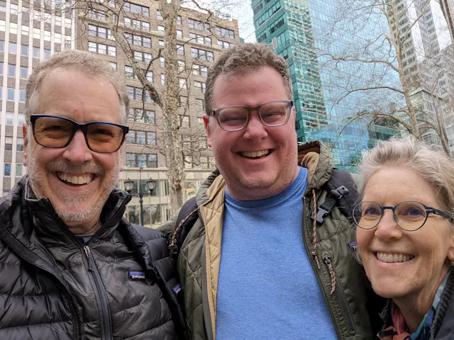 Three people taking a selfie outdoors in front of city buildings, wearing winter coats.