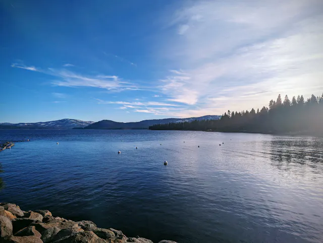 Calm mountain lake at dusk with pine trees and mountains reflected in the still water.