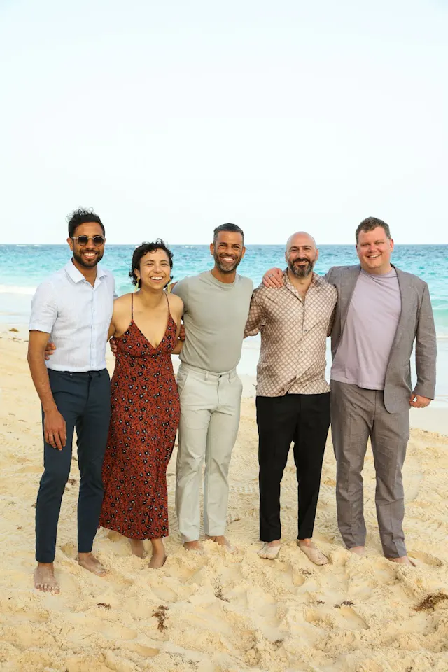 Five people posing together on a sandy beach with the ocean behind them.