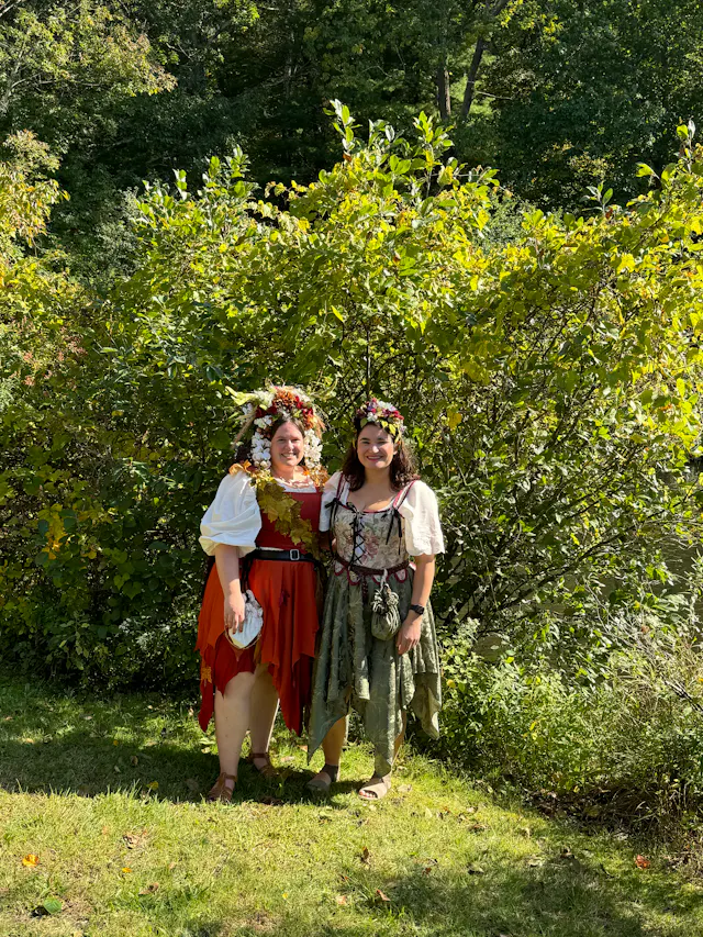 Two women in Renaissance faire costumes standing in front of green trees.