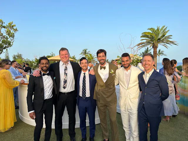 Six men in formal attire posing together at an outdoor wedding reception with palm trees.