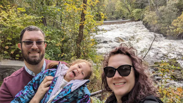 A man, woman, and young child taking a selfie in front of a rushing waterfall with autumn foliage.