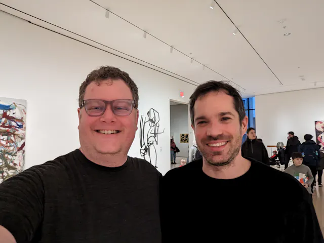 Two men taking a selfie inside an art museum with large paintings visible on the walls behind them.