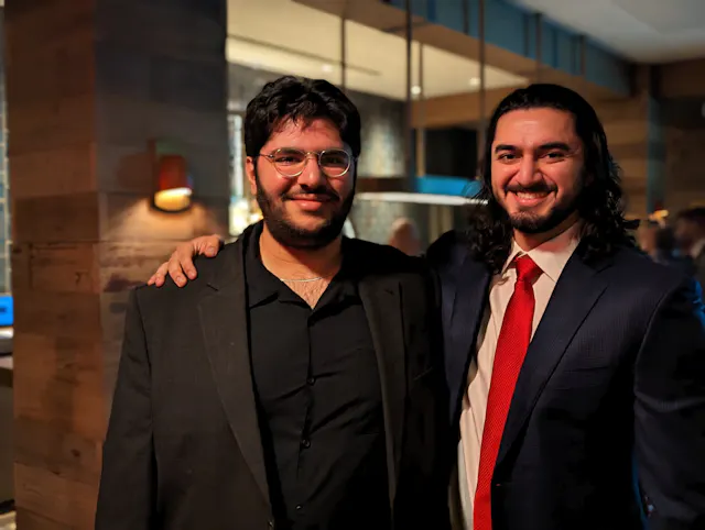 Two men posing together at a formal event, one wearing a black suit and the other a red tie.