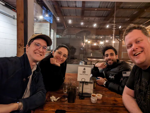 Four people smiling at a restaurant table with coffee cups in a warmly lit space.