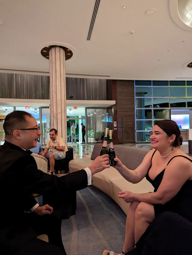 Two people in formal attire sitting in a hotel lobby, toasting with drinks.