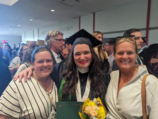 Three people at a graduation ceremony, the center person in cap and gown holding a bouquet of flowers.