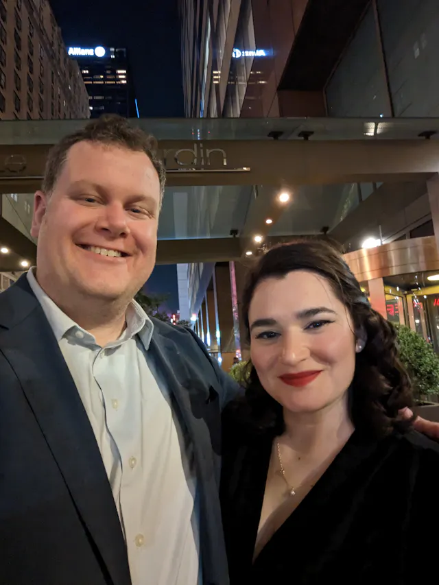 A couple taking a night selfie in front of a city hotel with lit signs behind them.