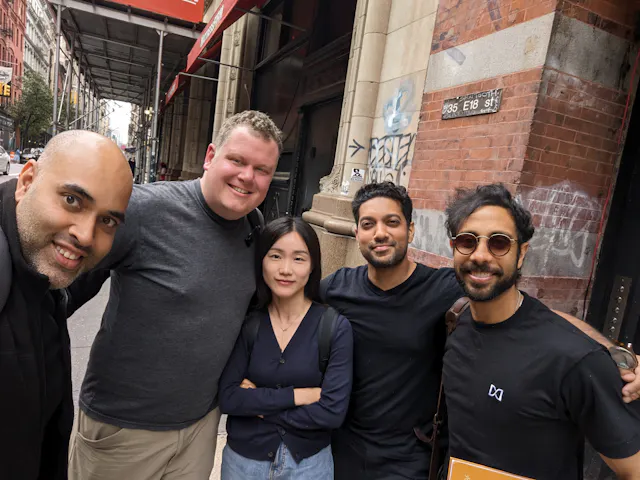 Five people taking a group selfie on a city sidewalk in front of brick buildings.