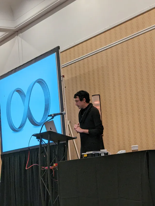 A presenter at a conference podium with a large projection screen showing an infinity symbol.
