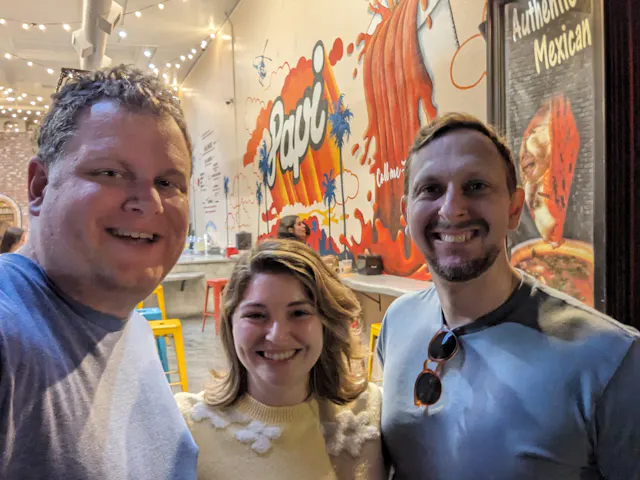 Three people smiling together in front of a colorful Mexican restaurant mural.
