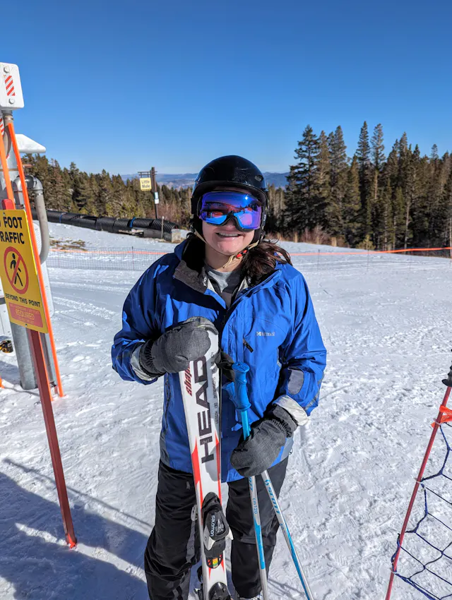 Person in ski gear and goggles smiling on a sunny snow-covered ski slope with pine trees.