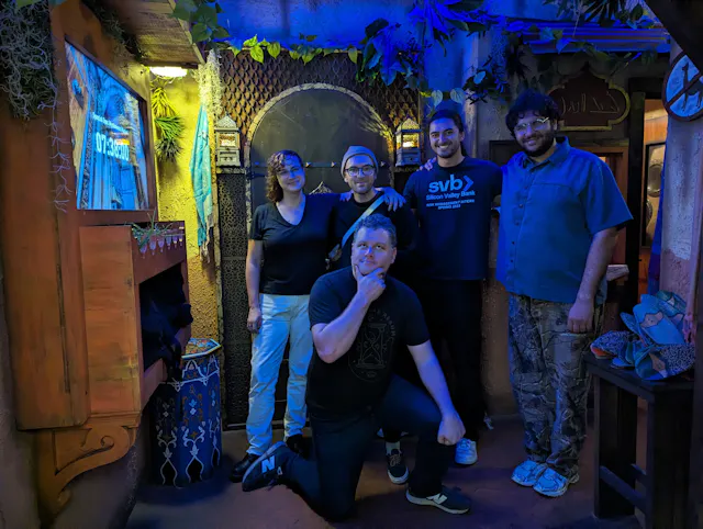 Five people posing in a dimly lit venue decorated with plants and colorful mood lighting.