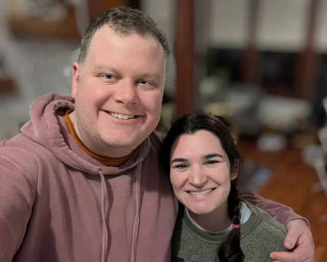 Two people smiling at the camera, one wearing a mauve hoodie and the other in a green sweater, indoors.