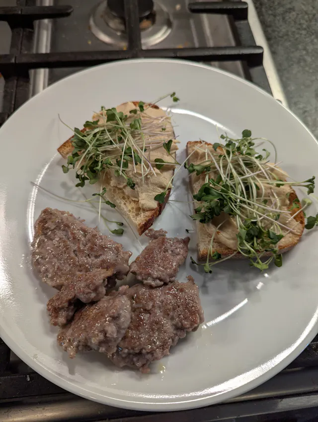 White plate with two slices of toasted sourdough topped with sprouts and hummus, accompanied by cooked breakfast sausage patties on the side.
