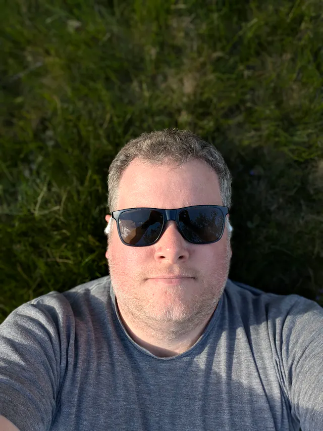 Selfie of a person wearing dark sunglasses and a gray striped shirt against a blurred green background.