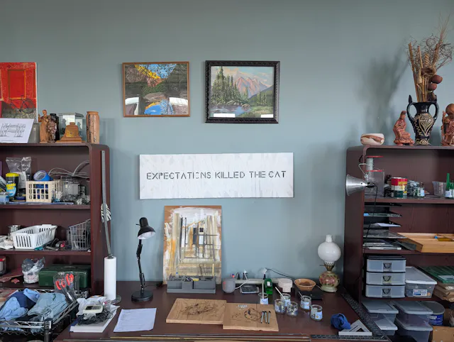 Wall display featuring framed artwork, a sign reading 'EXPECTING KILLED THE CAT,' shelves with objects, and a desk.