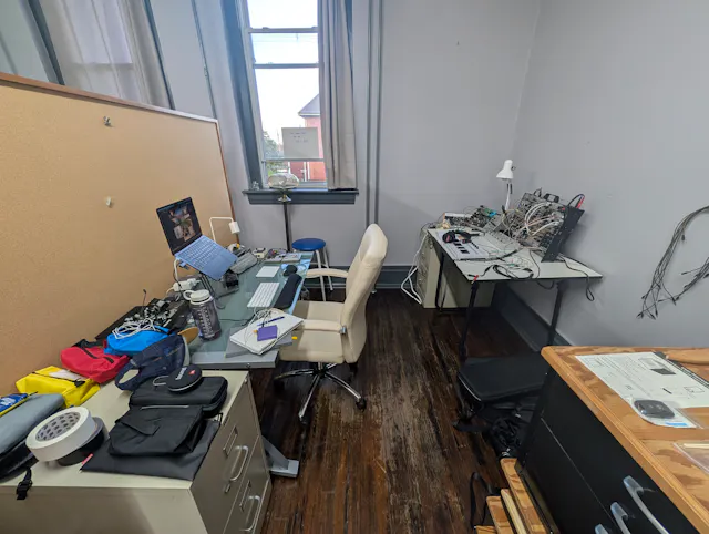 Cluttered studio workspace with colorful clothing, electronics, and supplies scattered on shelves and the floor.