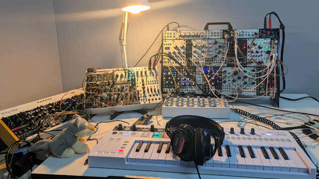 Overhead view of a white MIDI keyboard and headphones in front of several packed Eurorack cases, patch cables, a small desk lamp, and a tiny plush figure on a white desk.