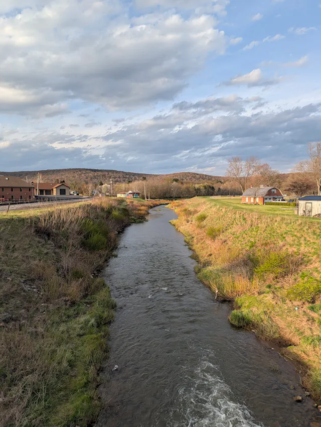 Wide stream flowing through pastoral landscape with green meadows, farm buildings, and tree-lined hills.