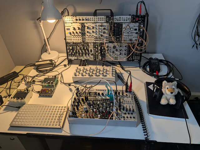 Modular synthesizer rack with multiple patch cables and control modules illuminated by desk lamp.