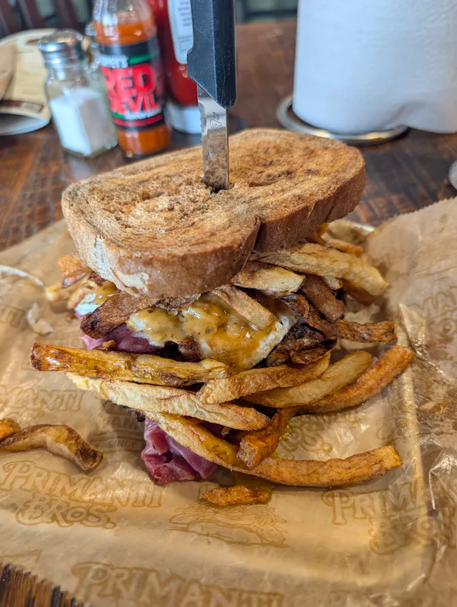 Tall stacked sandwich on Primanti-branded paper with a knife through the top, filled with crisp fries, sliced deli meat, and an egg, on a wooden table with condiments in the background.