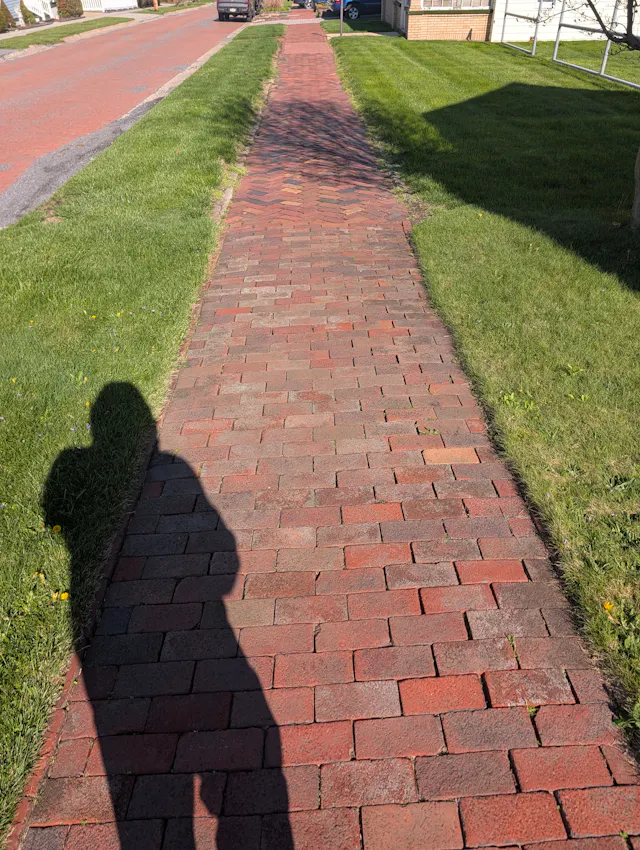 Long red-brick residential sidewalk receding into the distance in bright sun, with trimmed grass strips and a photographer's shadow in the foreground.