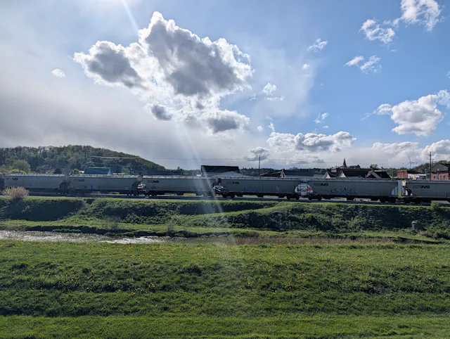 Long line of grey Norfolk Southern hopper cars on a track beside a river, green field with small yellow flowers, and a town with a church steeple under a partly cloudy sky.