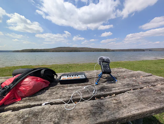 Field recording gear with a portable synthesizer, handheld recorder on a small tripod, and earphones on a weathered picnic table beside a lake and treeline.