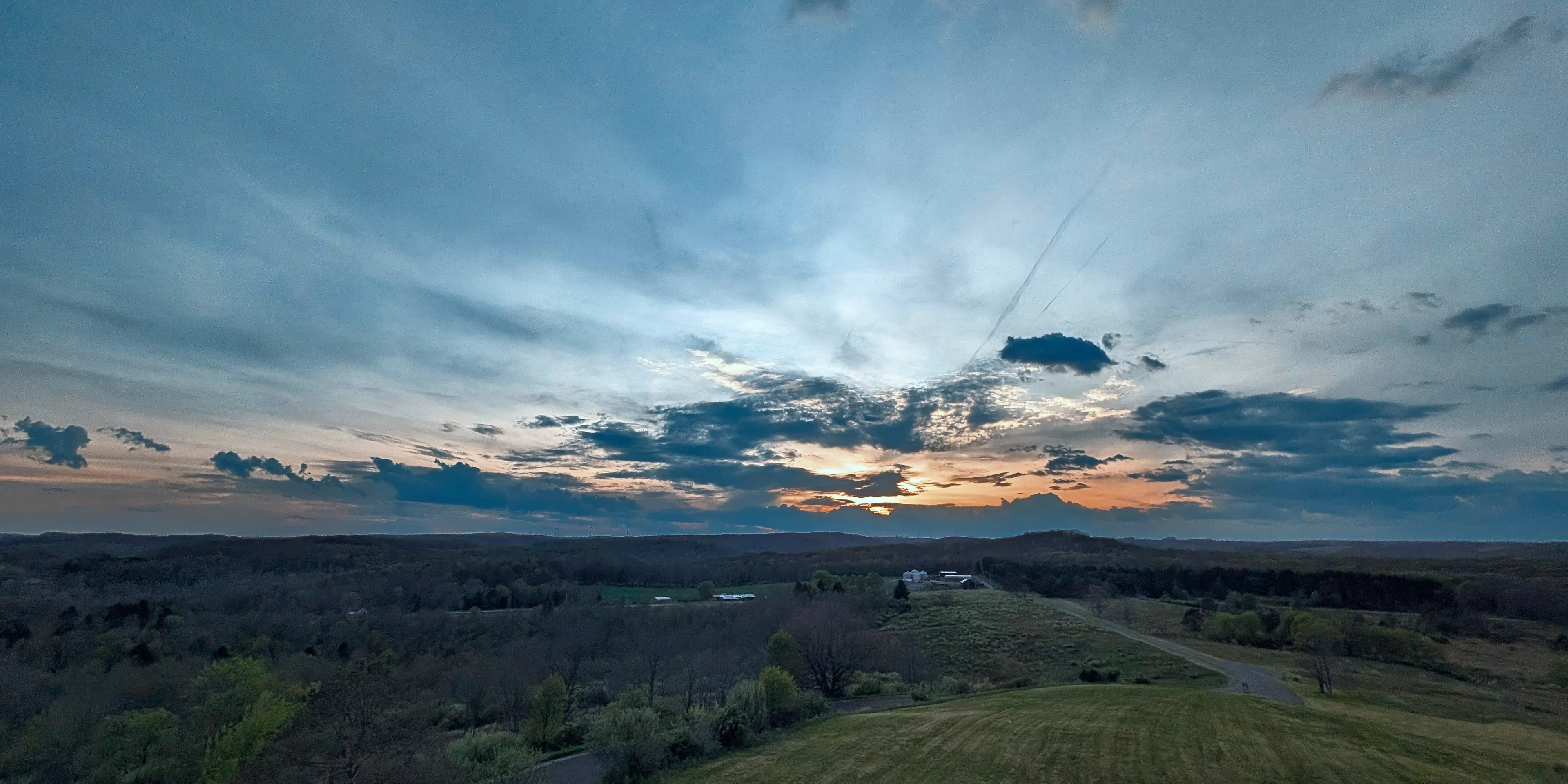 Rural scene at sunset with a deep blue sky fading to orange at the horizon, scattered clouds, a winding road, and a cluster of white farm buildings among tree-covered rolling hills.