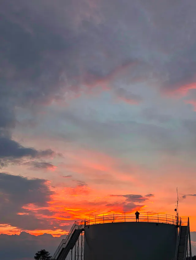 Silhouette of a person on the railed top of a large round industrial storage tank, with colorful orange, pink, and purple clouds in the evening sky above.