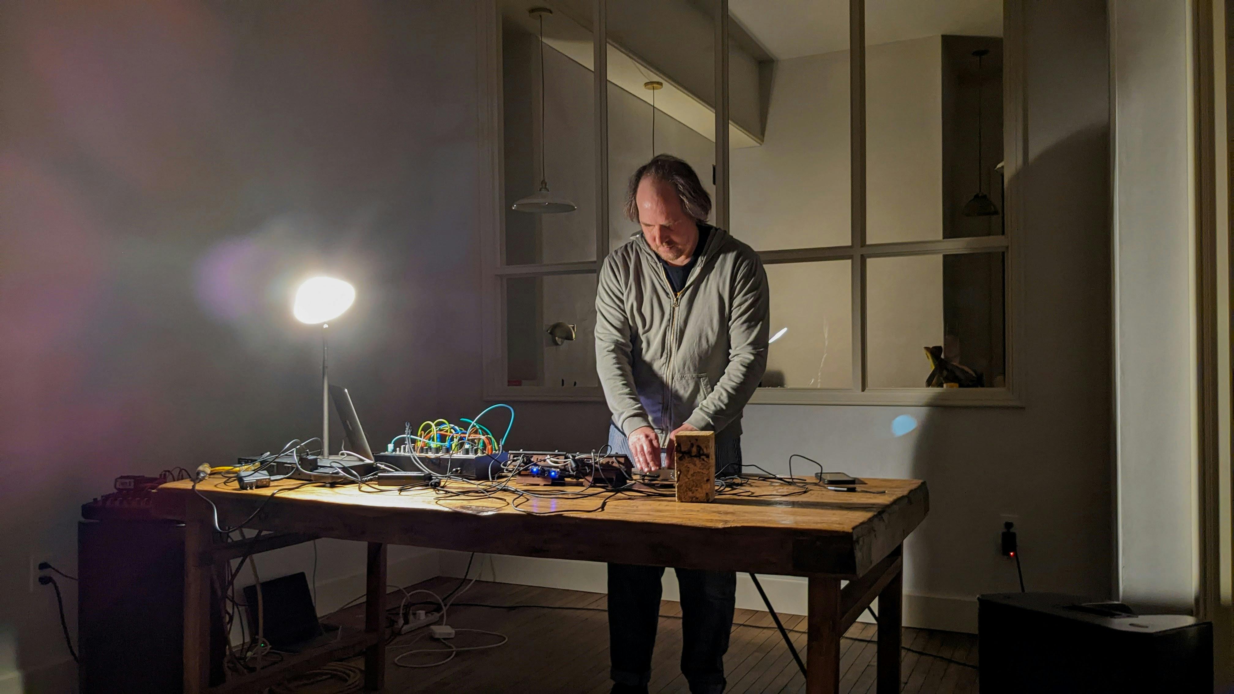 A man operates electronic music equipment on a wooden table in a room.