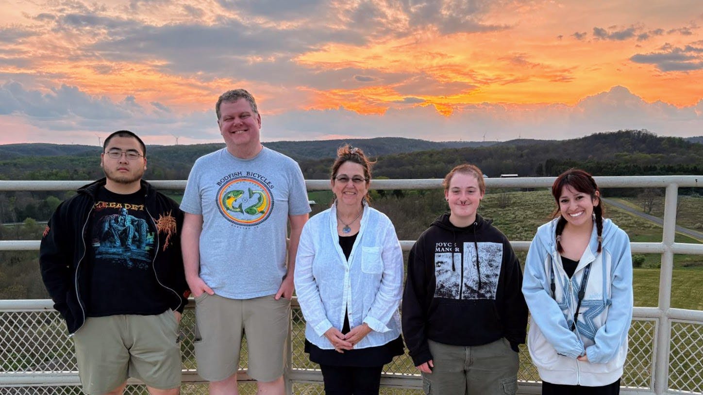 Five people stand on an overlook with rolling hills and a sunset behind them.