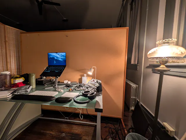 A studio desk lit by a small lamp and an antique globe lamp, holding a laptop on a stand, headphones, audio gear, and a wrist rest, with a tall cork divider behind it