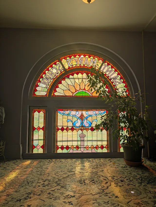 Sunlight pouring through a tall arched stained glass window labeled 'Saint Mary's School' (read in mirror image from inside), with colorful floral and geometric panels