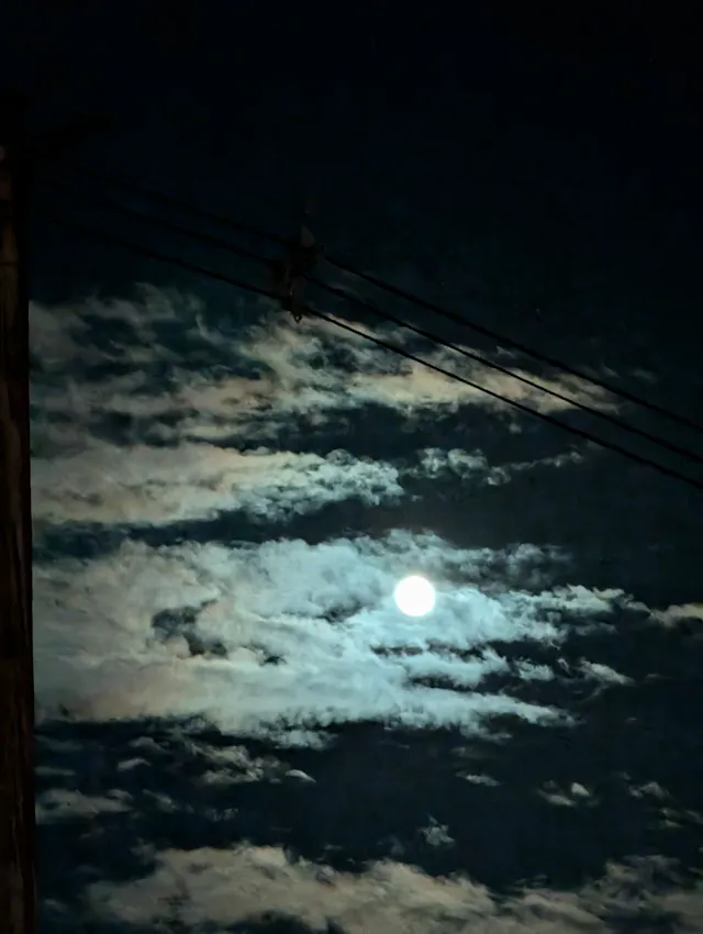 A bright full moon glowing through scattered clouds at night, with utility wires crossing the upper part of the sky