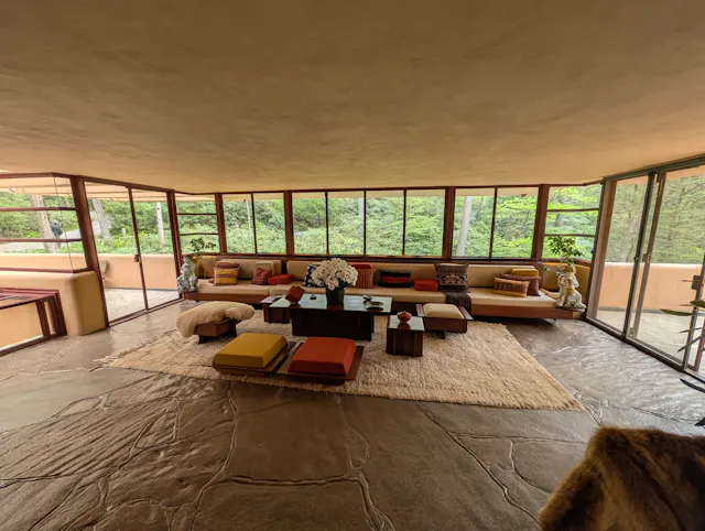 Fallingwater's living room with built-in low couches, leather ottomans on a wool rug, a flagstone floor, and ribbon windows looking out into the forest