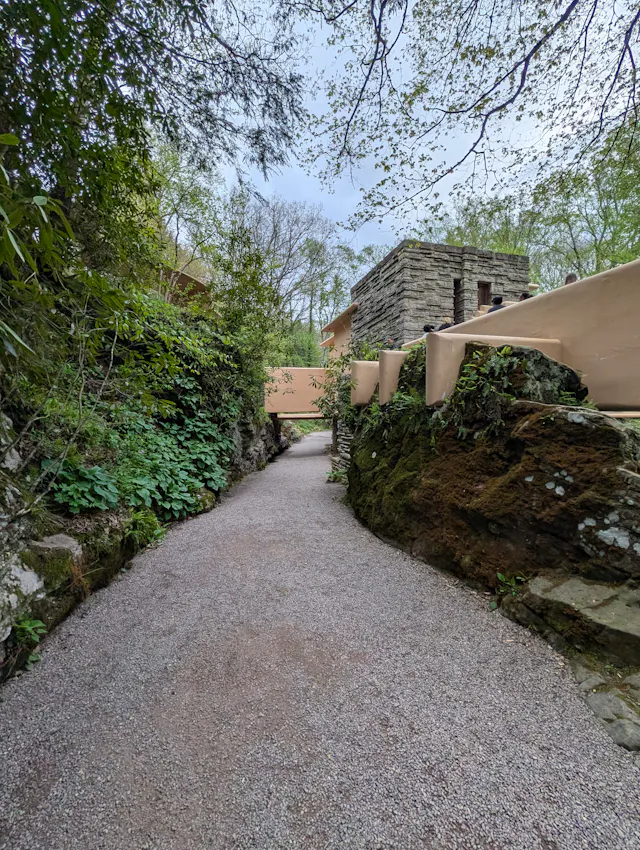 A gravel walking path leading toward Fallingwater between mossy boulders and a low concrete wall