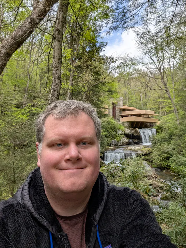 Selfie of Nat in a black fleece jacket smiling in front of Frank Lloyd Wright's Fallingwater and its waterfalls